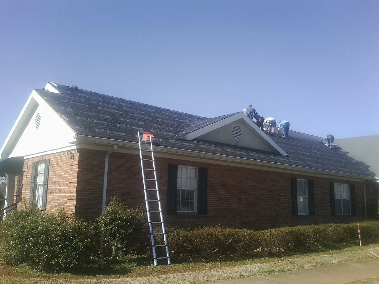 Crew tearing shingles off a brick ranch roof with a ladder set — Emergency Roof Repair work by our crew in Cooperstown